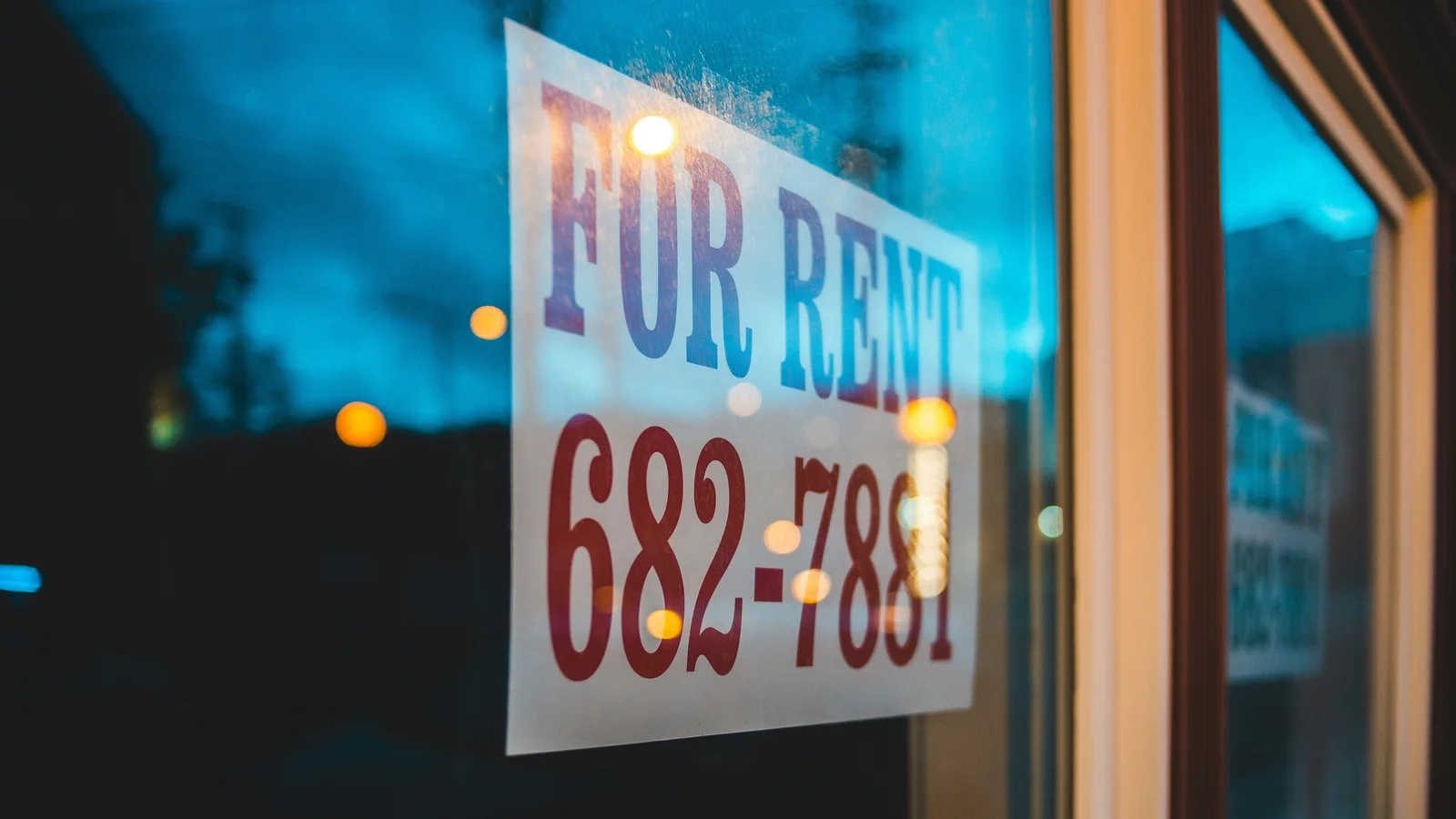 A "For Rent" sign in a shop window at dusk.